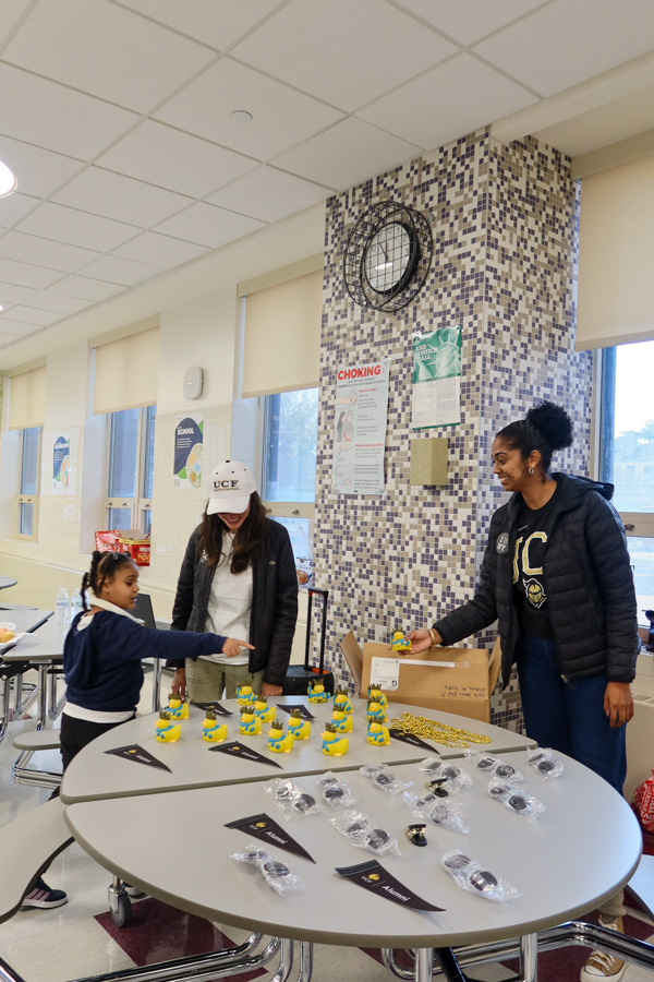 Girl in a school uniform points at yellow Minion figurines lined up on a round table during a school event; two adults organize items from a box nearby.