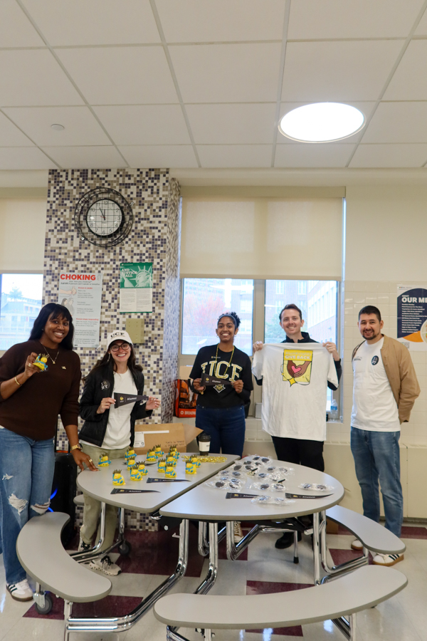 Five diverse people stand behind a table with small yellow-and-green promotional items in a school cafeteria, smiling at the camera.