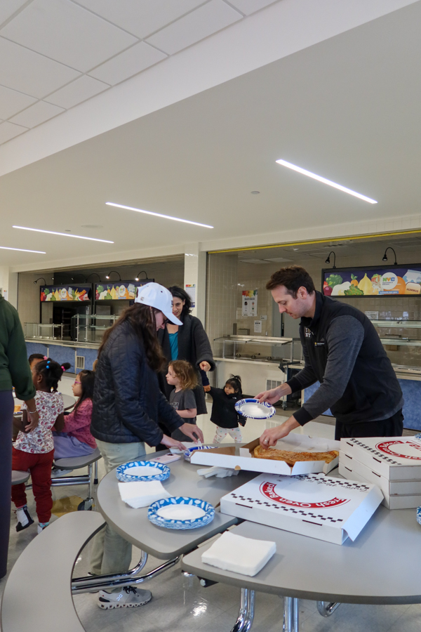 People gather around a round cafeteria table while a man hands out pizza slices from an open box with pizza boxes and plates nearby.
