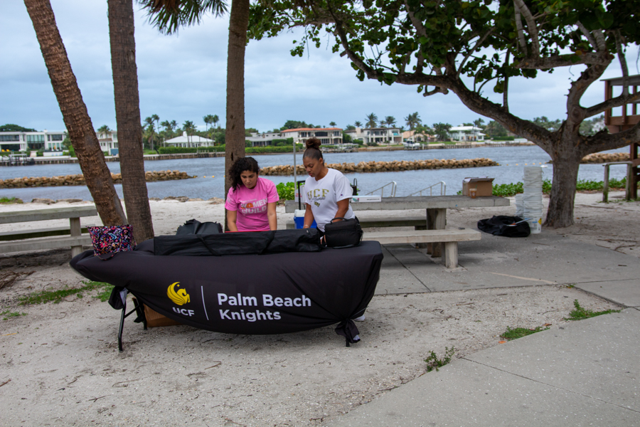 Two people setting up a table with a Palm Beach Knights banner by a waterfront park with palm trees and benches nearby.