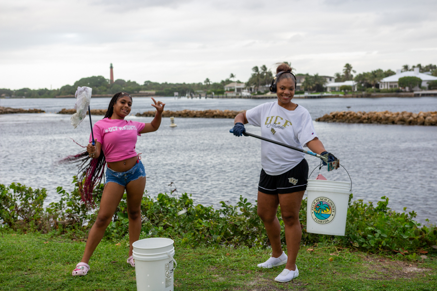Two young women cleaning a shoreline: one with a net and pink shirt, the other with a bucket and long grabber by the water.