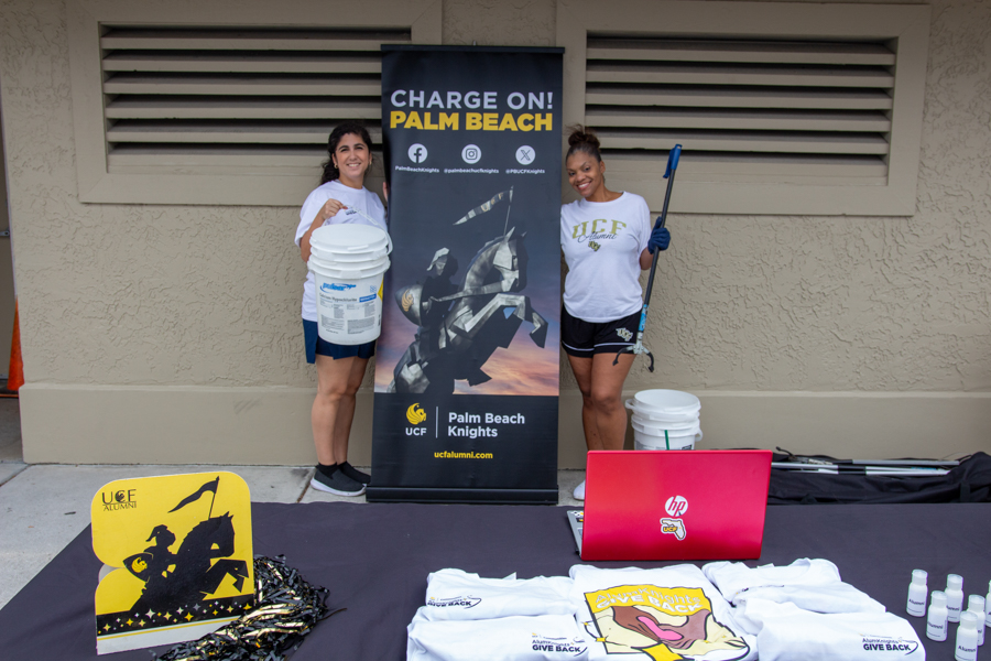 Two women standing beside a vertical banner that reads 'Charge On! Palm Beach Knights' at an alumni event, holding cleaning buckets and tools.