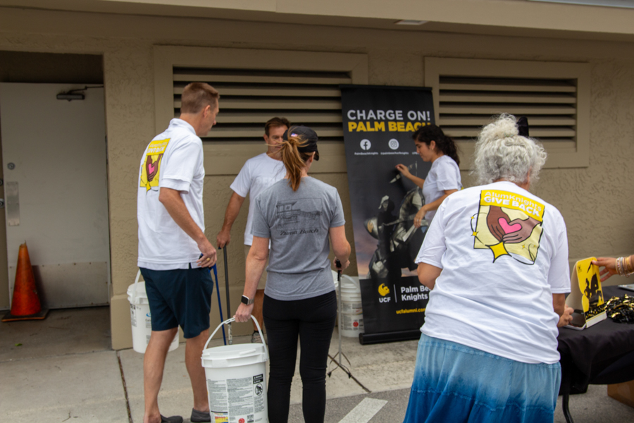 Group of volunteers in white shirts gathered near a promotional banner at an outdoor charity event, with white buckets in hand.