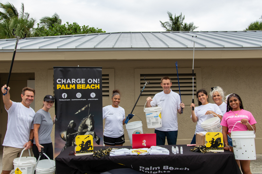 Eight volunteers stand behind a table with buckets and gear at a 'Charge On! Palm Beach' event banner, ready to fundraise or clean up.