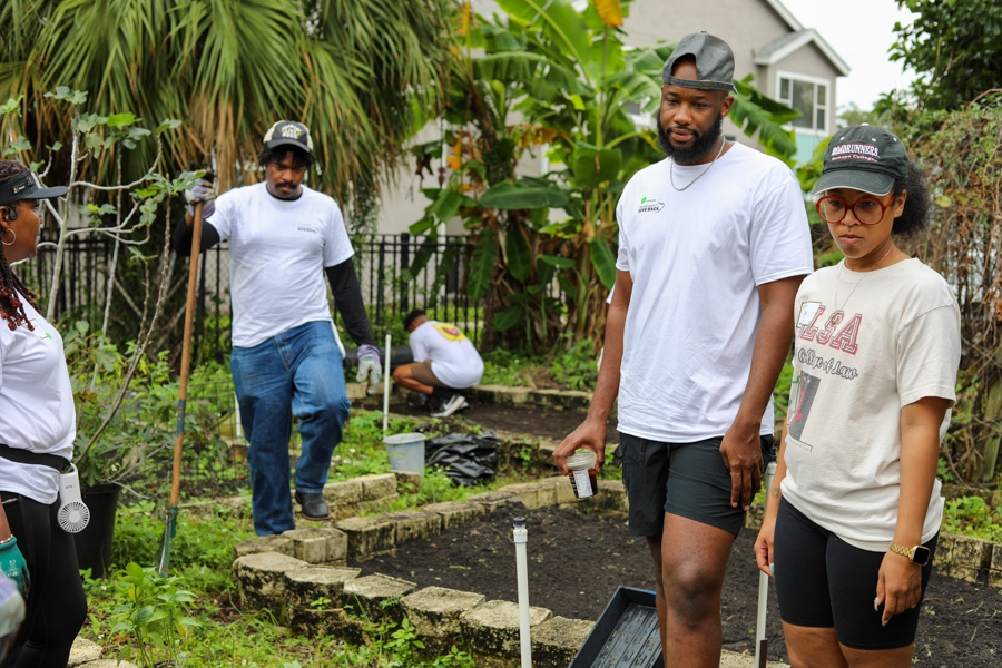 Group of volunteers in a backyard garden tending raised beds and plants, some with shovels and tools in hand in a sunny yard.
