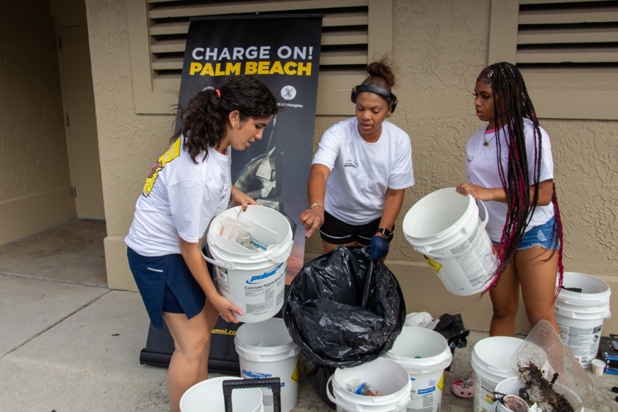 Three volunteers gather trash for a beach cleanup, placing items into white buckets beside a banner that reads 'Charge On! Palm Beach'.