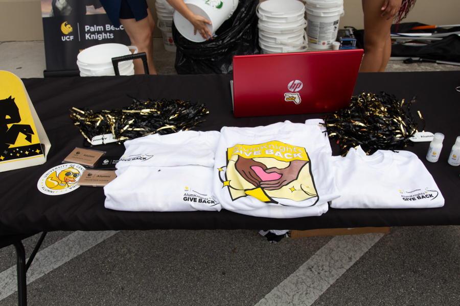 Table with giveaway shirts and event props for a student charity drive; shirts read 'Give Back' and feature a cartoon mouth logo, plus a laptop and pom-poms on the table.