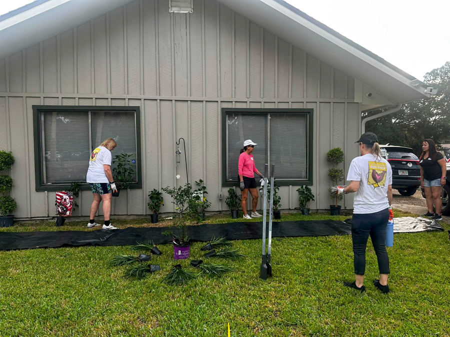 Group of volunteers planting shrubs along the front of a beige house with black ground cover mats and potted plants nearby.