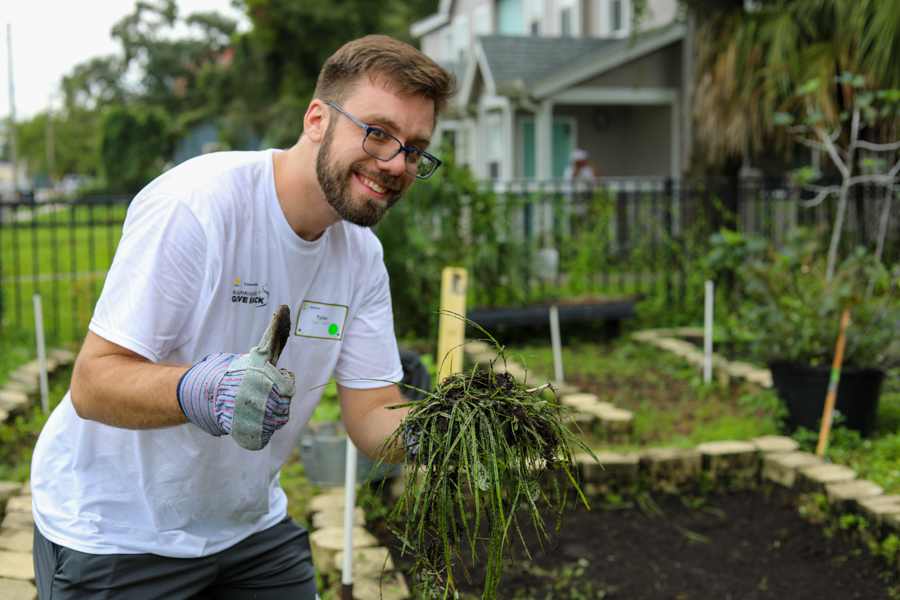 Smiling man with glasses and gloves giving a thumbs up while holding a clump of wet plant roots in a garden yard.