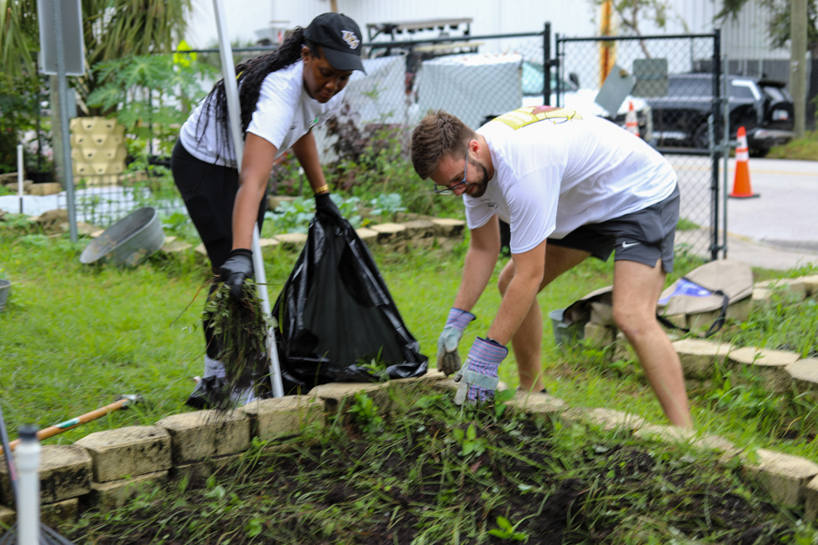Two volunteers weed a garden bed, placing clippings into a trash bag nearby.