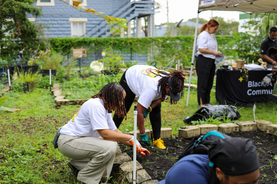 Volunteers in white shirts kneel and plant in a raised garden bed at a community garden event, with others and a canopy in the background.