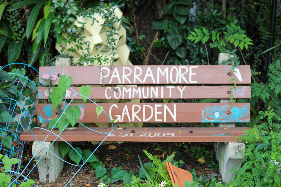 Wooden bench with a painted sign reading 'PARRAMORE COMMUNITY GARDEN', surrounded by lush green plants and vines