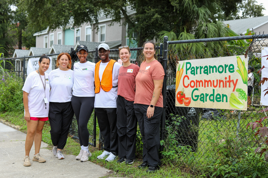 Six volunteers pose together in front of a chain-link fence with a Parramore Community Garden banner in the background.