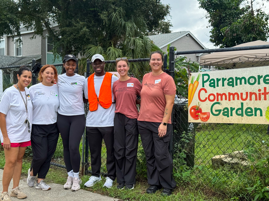 Six volunteers pose together in front of a Parramore Community Garden banner, smiling outdoors. (group photo)
