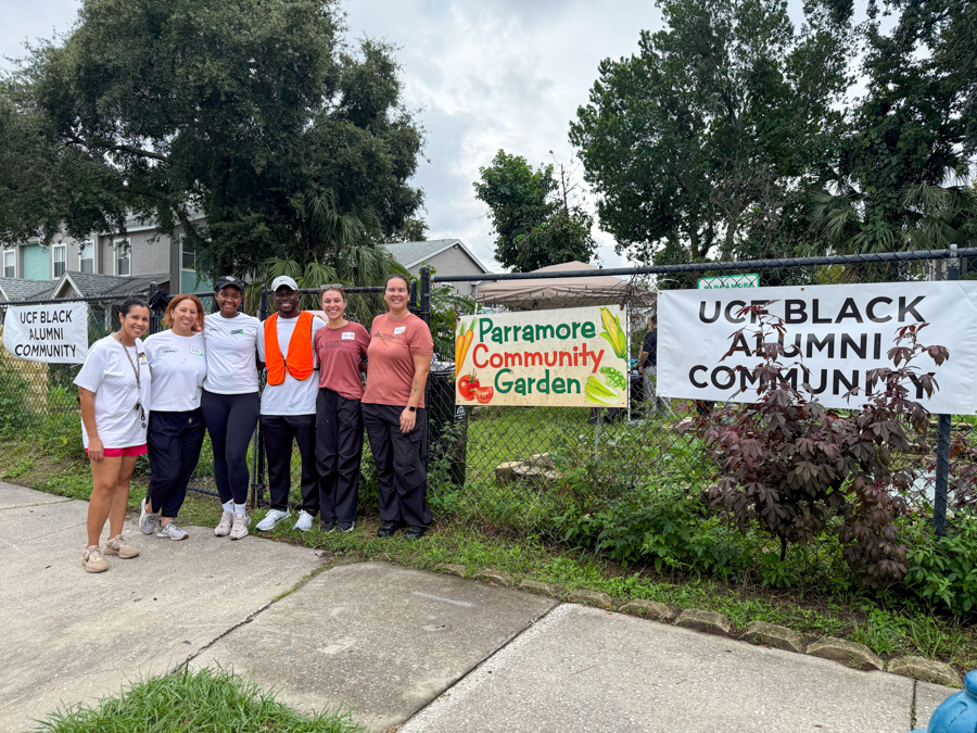 Group of volunteers posing by a chain-link fence with Parramore Community Garden and UCF Black Alumni Community banners behind them during a sunny day.