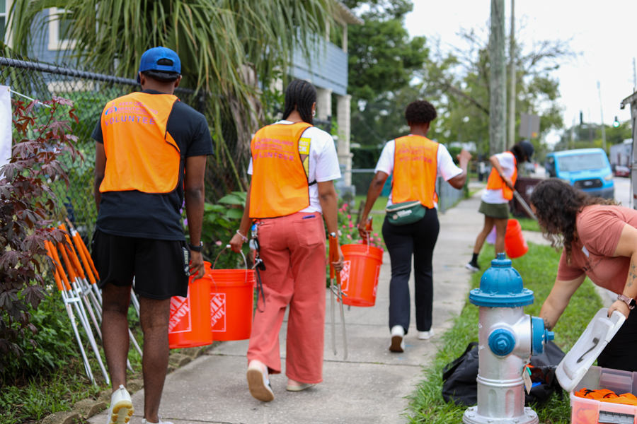 Neighborhood cleanup volunteers in orange safety vests walk along a sidewalk carrying orange buckets and tools.