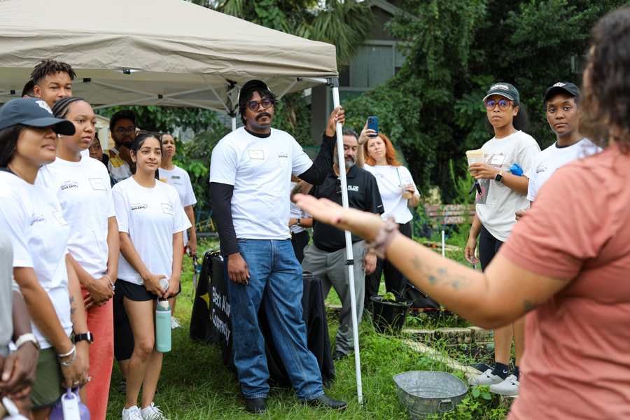 Group of volunteers under a canopy listening to a speaker at an outdoor garden workshop at a community event.