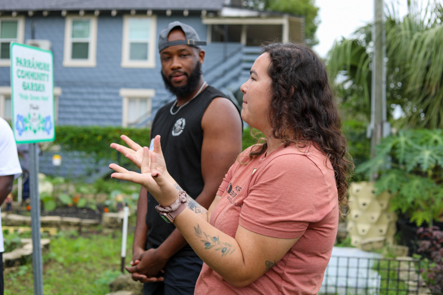 Two adults converse in a community garden; the woman in a peach shirt gestures with her hands while a man in a black tank top stands nearby, with a sign for the garden in the background.