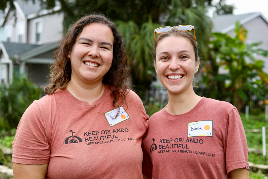 Two smiling volunteers in coral Keep Orlando Beautiful shirts pose together in a residential garden.