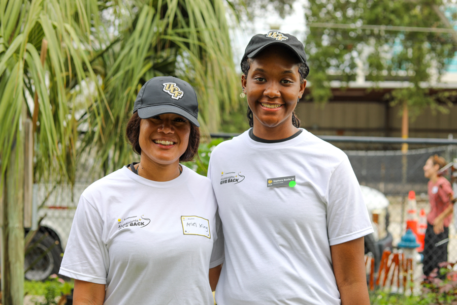 Two smiling women in matching white T-shirts and dark caps with logos, posing outdoors at a community event with greenery in the background and a chain-link fence behind them.