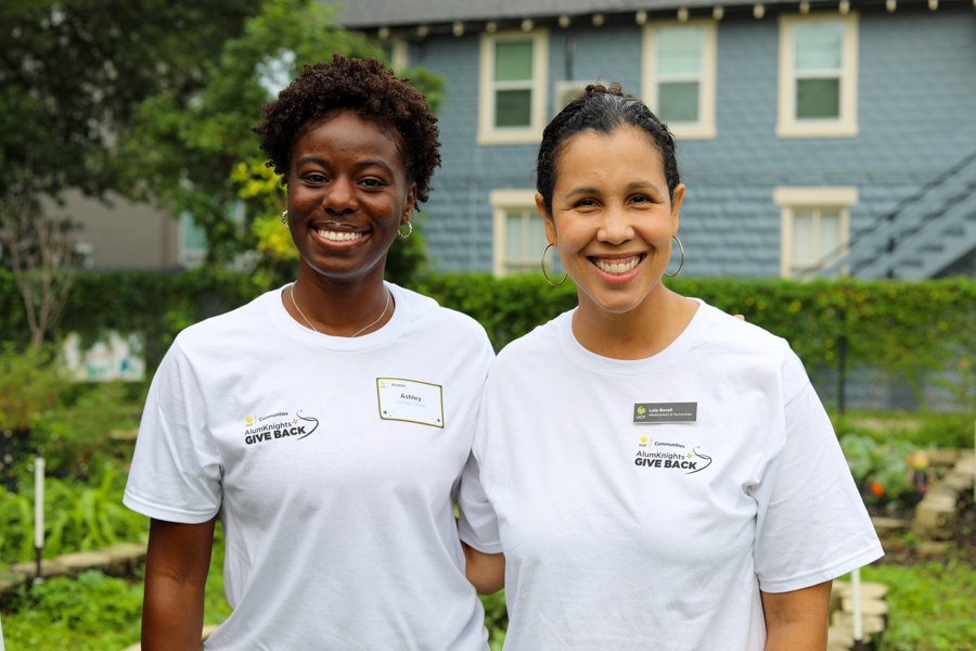 Two smiling women volunteers in white 'Give Back' shirts pose outdoors in a garden with a blue house in the background, arms around each other.