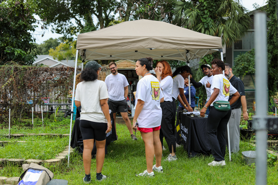 Group of volunteers under a beige canopy at an outdoor event, standing around a table with merchandise or supplies and talking to attendees in a grassy yard.