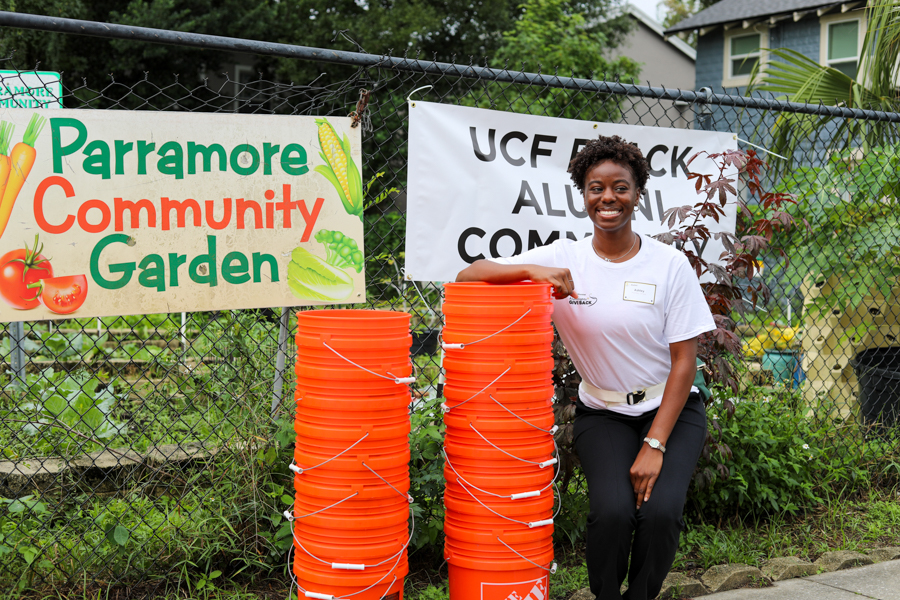 Smiling woman in a white T-shirt leans on tall orange buckets in front of a chain-link fence, with banners reading 'Parramore Community Garden' and 'UCF Alumni' behind her among garden plants.