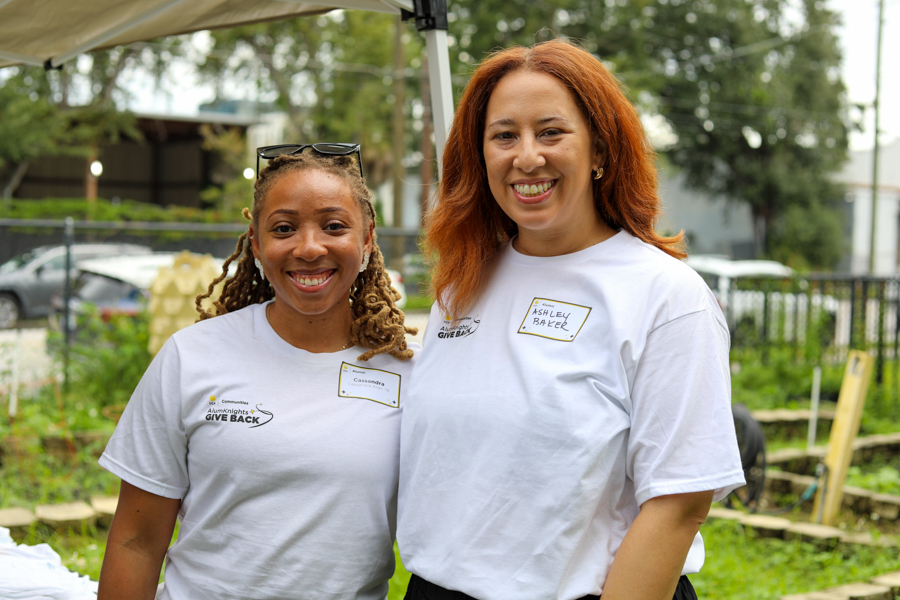 Two smiling women volunteers in white T-shirts at an outdoor community garden event.