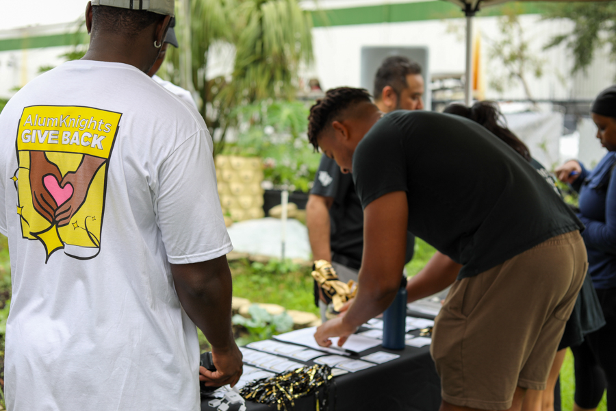 Volunteer event outdoors; person in white shirt with AlumKnights GIVE BACK logo watches others arrange papers at a registration table outside under a canopy.