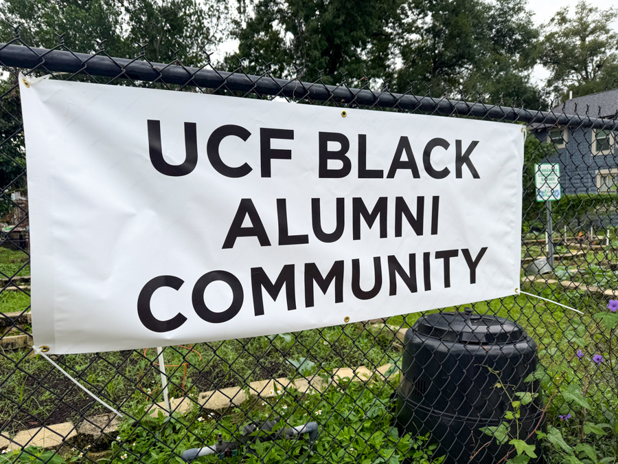 White banner on a chain-link fence reading 'UCF BLACK ALUMNI COMMUNITY' in a garden area, with greenery around it.