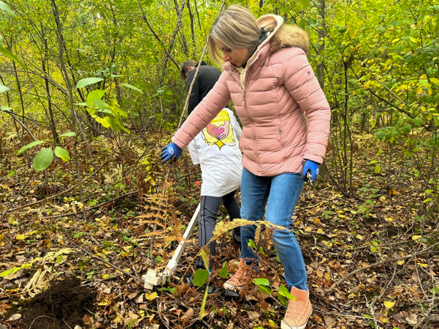 Woman in a pink puffer jacket plants a sapling in a forest with a child nearby, wearing gloves and using a shovel in loose leaves.