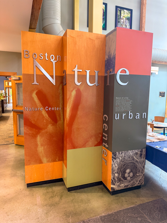 Inside a nature center with tall orange display pillars and large white text, including 'Boston Nature Center' and adjacent 'urban' panel.