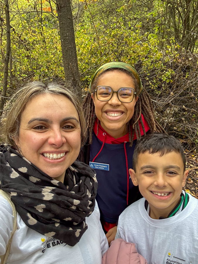 Three people posing for a selfie in a forest, all smiling at the camera with autumn leaves in the background.
