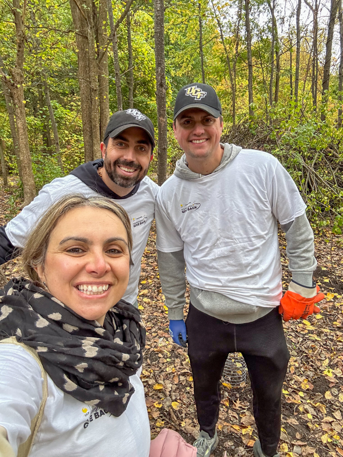 Smiling woman takes a selfie with two men in a wooded fall setting; all wear white event shirts and gloves, likely volunteering outdoors.