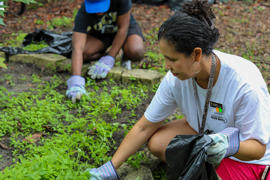 Volunteer woman in a white T-shirt weeds a garden bed, wearing gloves, with another helper kneeling in the background.