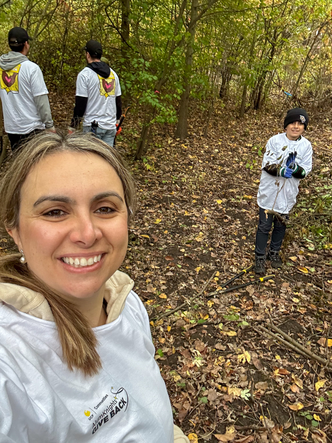 Smiling woman in a forest taking a selfie during a volunteer cleanup; volunteers in white shirts work behind her on a leaf-strewn trail.