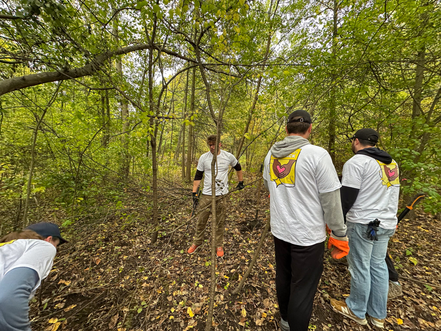 Group of volunteers in matching white shirts with a heart logo clearing brush in a wooded area; some wear gloves and use garden tools.