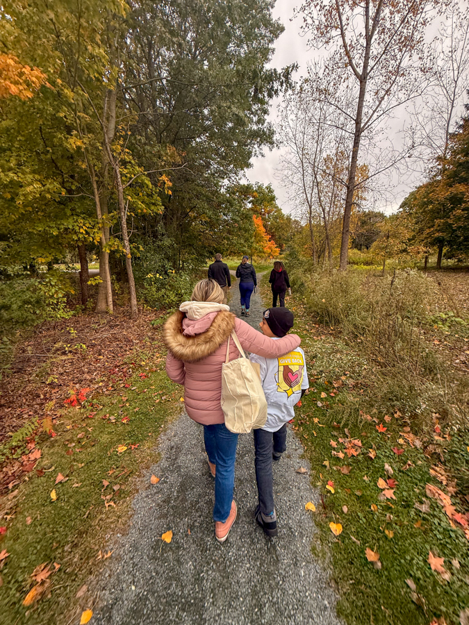 Woman in a pink jacket with a fur hood walks arm-in-arm with a child in a gray hoodie along a leaf-strewn park path in autumn ground cover and colorful trees ahead, others visible in distance.