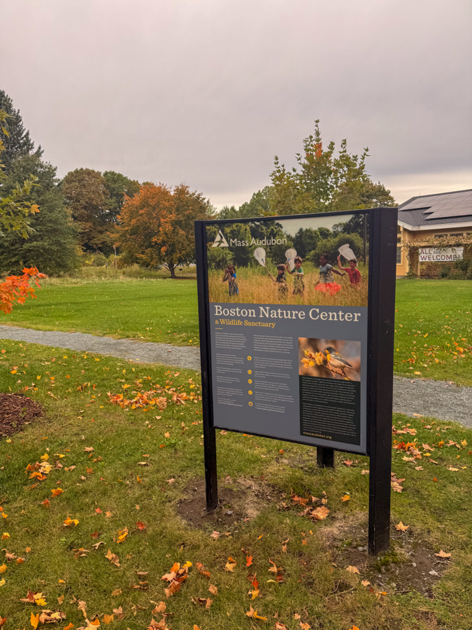 Signboard at Boston Nature Center (Mass Audubon) with a photo of children in a field and text about the sanctuary.