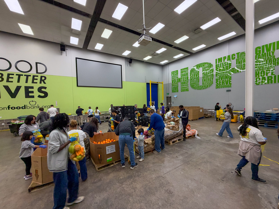 Volunteers sort and box fresh produce in a large food bank warehouse with a green wall and slogans above.