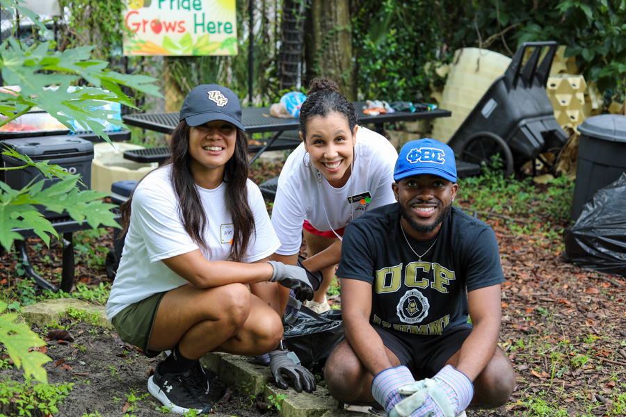 Three volunteers crouch and smile in a garden cleanup area with plants and tools around them with a sign in the background.