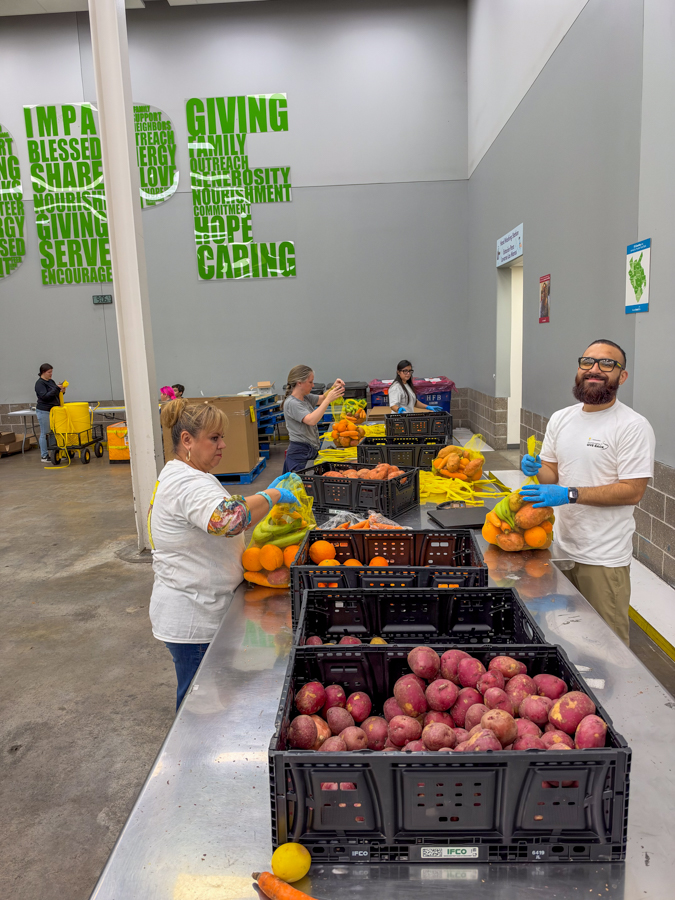 Volunteers bagging fresh produce at a long table in a food bank, with crates of oranges and apples and a green wall art in the background.