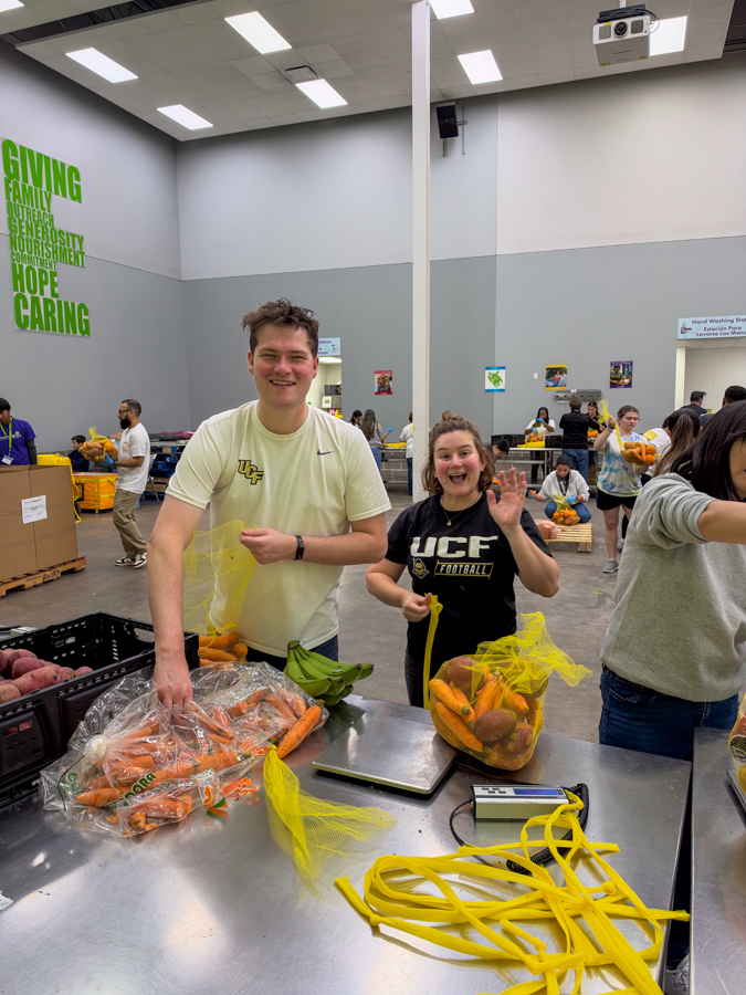 Two volunteers smile at a table sorting bags of carrots and other produce at a large indoor food drive event.