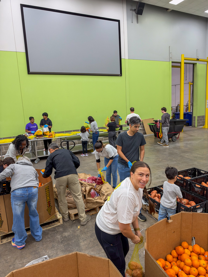 Group of volunteers sorting and bagging Produce in a large indoor facility, with crates of onions and oranges in the foreground and a bright green wall in the background.