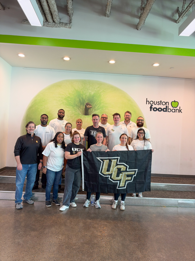 Group of diverse people standing on a stage-like platform, holding a UC flag inside a Houston Food Bank facility.