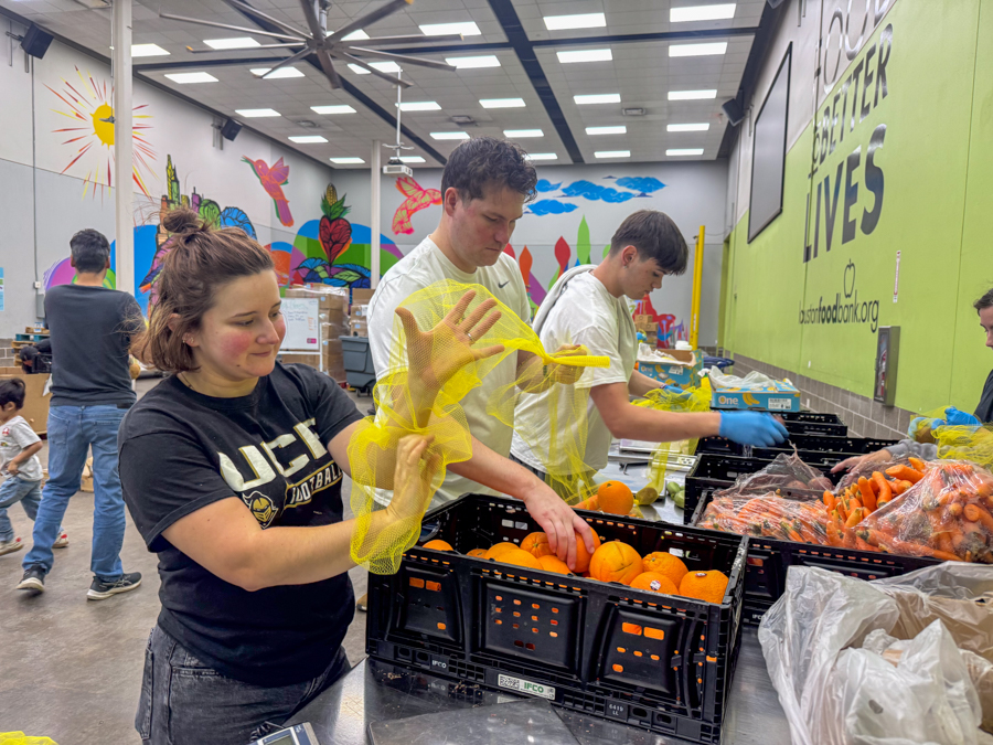 Volunteers in a bright warehouse sort oranges into crates at a food bank packing station.