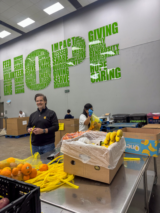 Volunteer in a black shirt packs fruit at a donation center; a woman in a mask sorts produce in the background under a large green 'GIVING' mural on the wall.