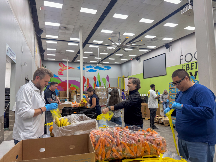 Volunteers wearing gloves bagging carrots and other produce at a food bank packing station in a bright warehouse.