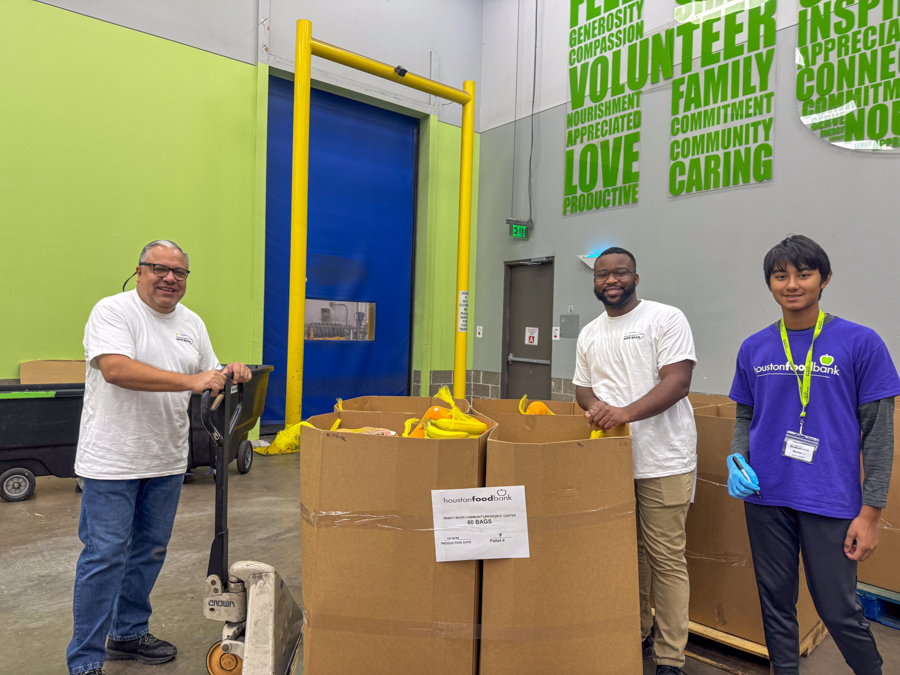 Three volunteers stand in a warehouse beside large cardboard bins filled with donated bags; a man on a pallet jack is on the left, another smiling man is center, and a young person in a purple Houston Food Bank shirt is on the right.
