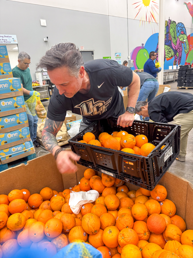 Volunteer lifts a crate of oranges into a large tub at a donation center, with others sorting in the background.